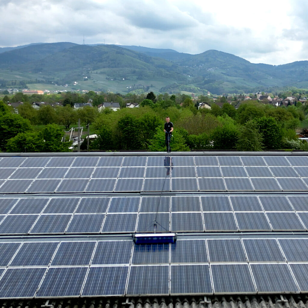 Photovoltaikreinigung-Bühl-Skyline-Solarninjas-01 Mitarbeiter reinigt Photovoltaikanlage auf Industriedach in Bühl mit Blick auf den Schwarzwald – Photovoltaikreinigung Bühl